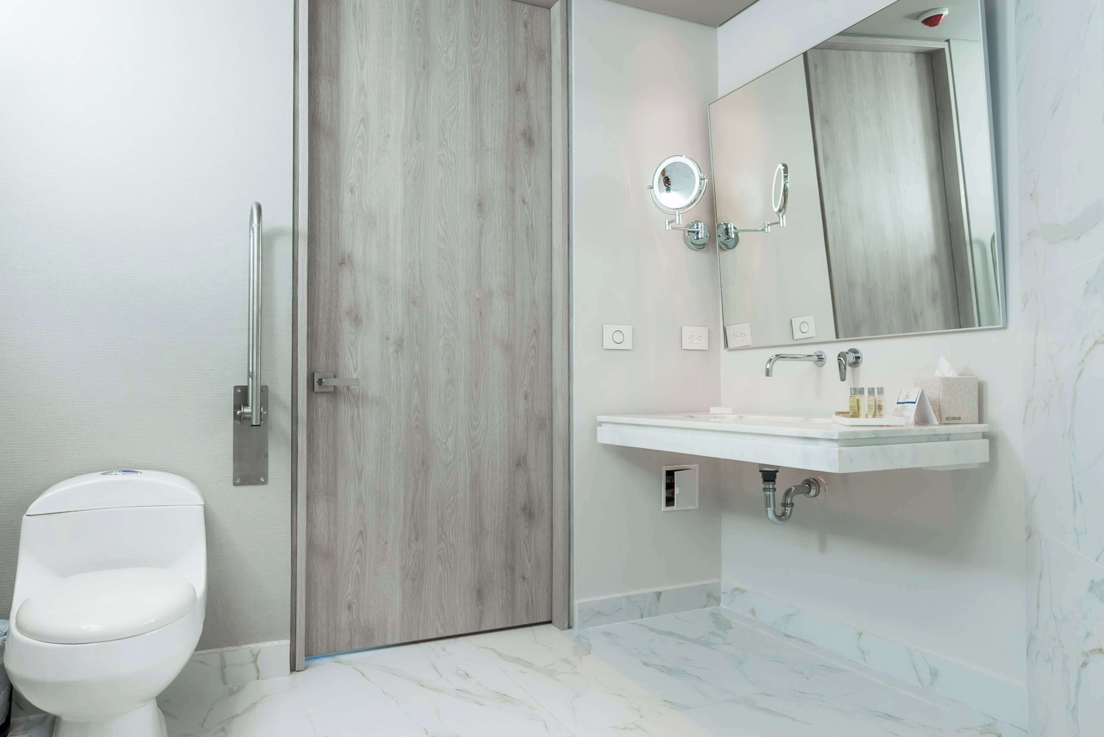 Un baño moderno con puerta de madera, inodoro blanco, lavamanos con espejo y suelo de mármol.
