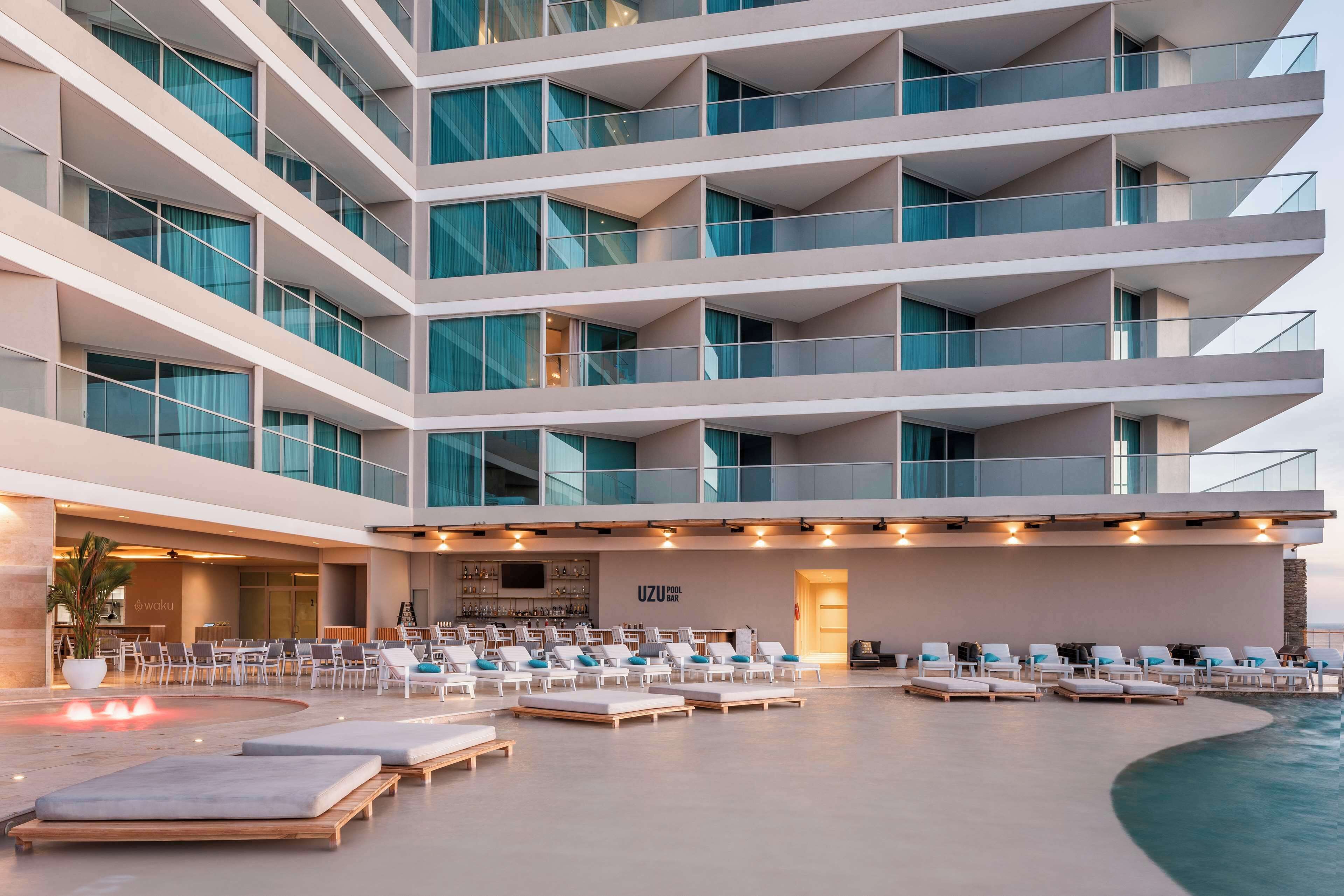 A modern hotel with multiple balconies, a pool area with sun loungers, and a bar/lounge area.