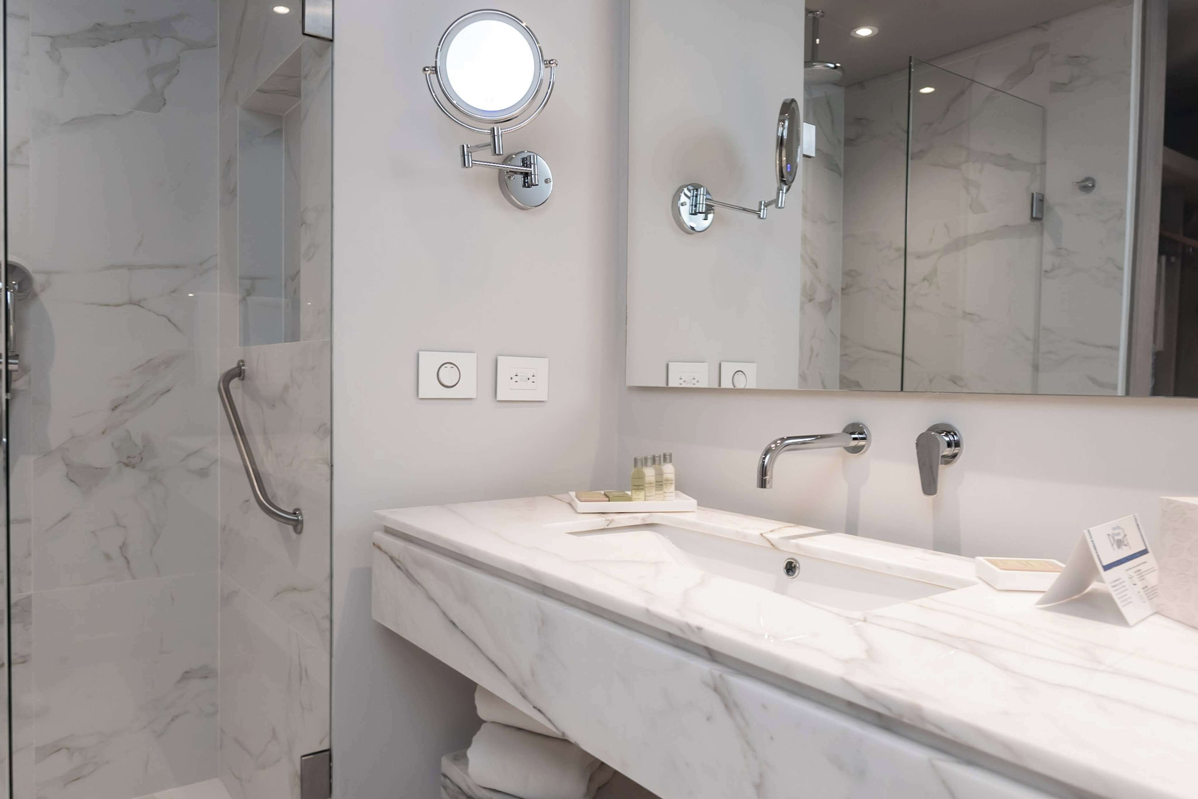 A modern bathroom with a marble vanity, a large mirror, a handheld showerhead, and a glass-enclosed shower.