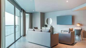 Una habitación de hotel moderna con un sofá grande, un sillón más pequeño, un televisor montado en la pared y un balcón con puertas correderas.
