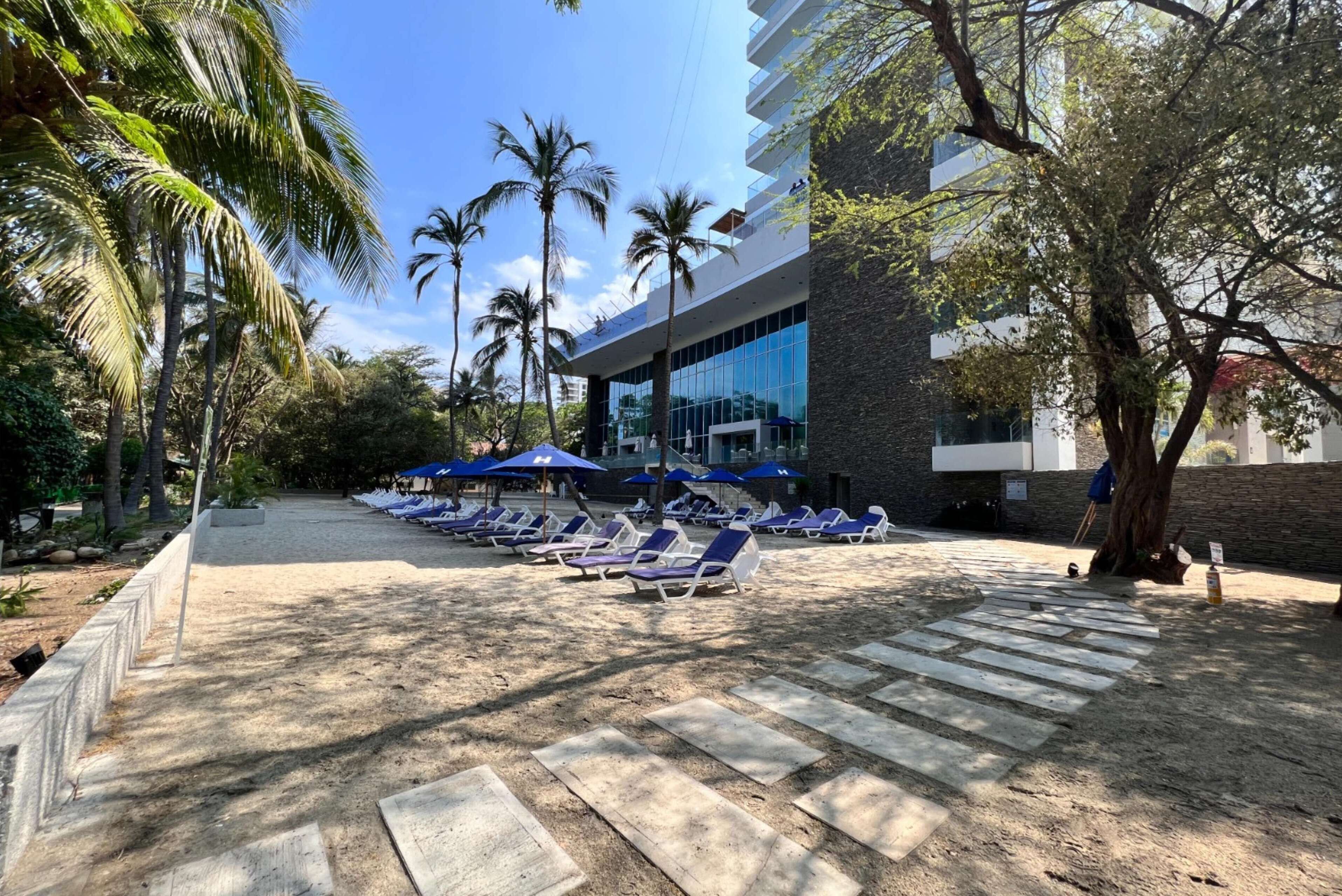 A poolside area with lounge chairs and umbrellas, palm trees, and a modern building with reflective glass windows.