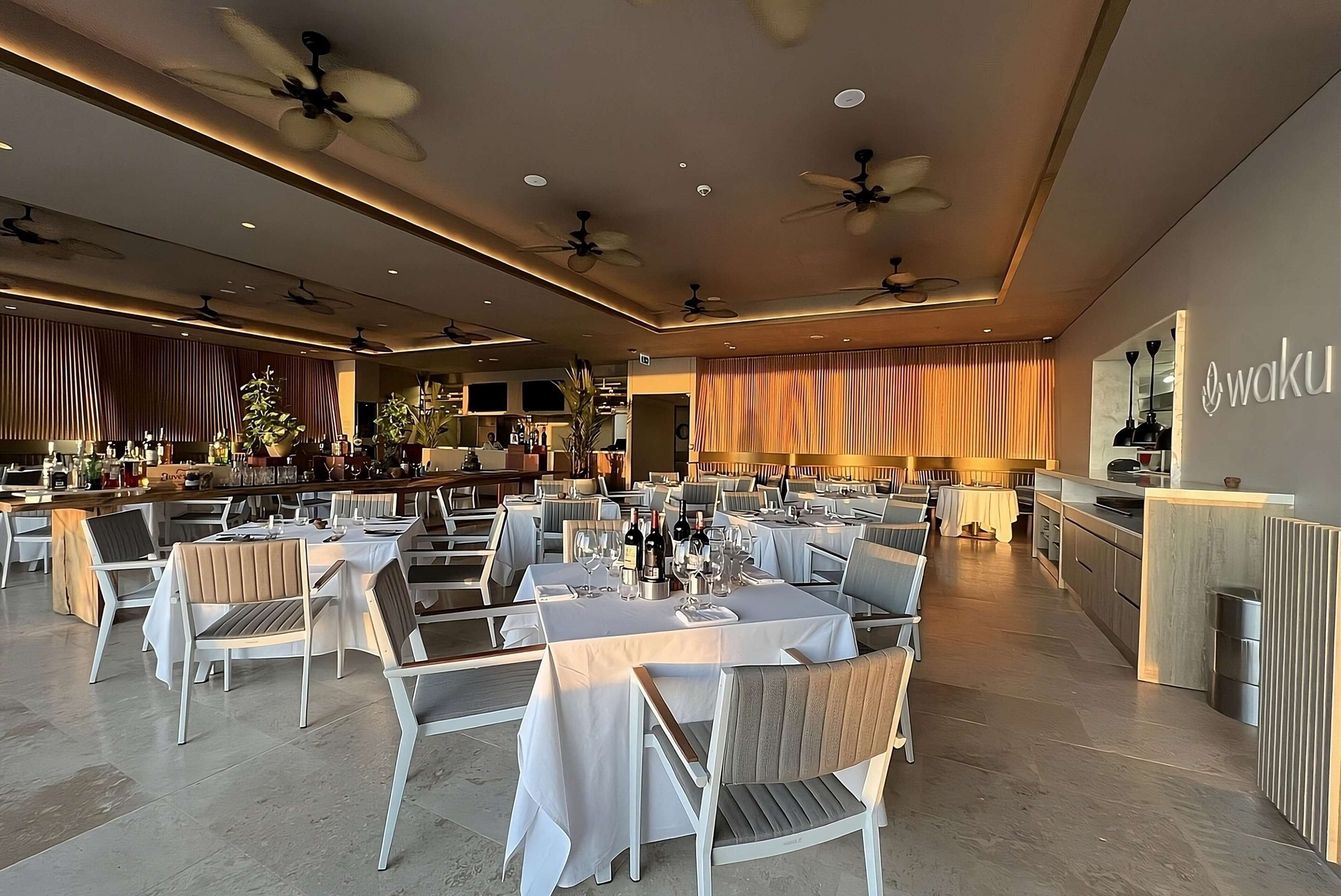 A spacious dining area with multiple tables set for guests, ceiling fans, and a bar area.