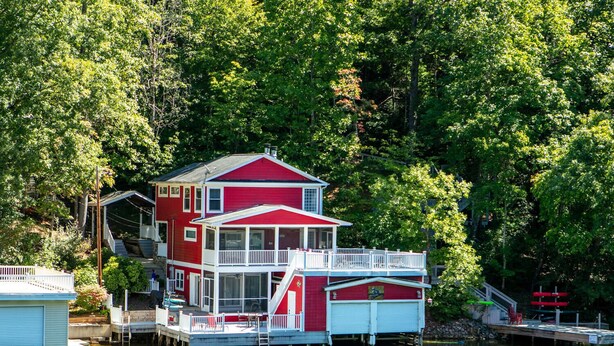 Una casa flotante roja y blanca con balcón, amarrada en un cuerpo de agua tranquilo rodeado de árboles verdes y frondosos.