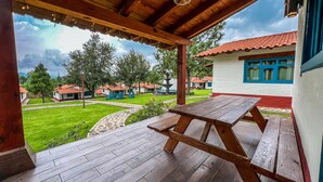 Un patio cubierto con una mesa y bancos de madera, con vistas a un césped cuidado y varios edificios con tejados de tejas rojas.