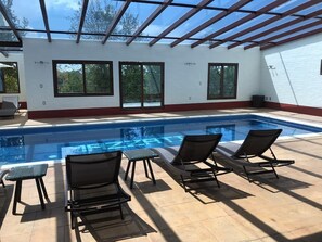 A swimming pool with four lounge chairs and a glass table, surrounded by large windows and a covered patio area.