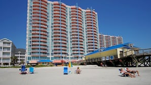 A high-rise hotel with multiple balconies, a beach area with sun loungers, and a pier structure. - 1st Rate Views & Amenities - Ritzy Oceanfront Family Resort w/pools, pier restaurant/bar in Ocean Dr (North Myrtle Beach)