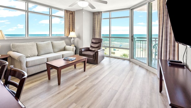 A spacious living room with a large window offering a view of the beach, a ceiling fan, and a leather armchair. - 1st Rate Views & Amenities - Ritzy Oceanfront Family Resort w/pools, pier restaurant/bar in Ocean Dr (North Myrtle Beach)