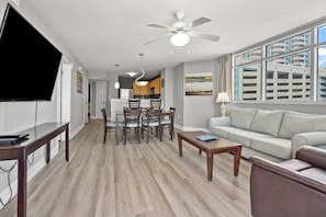 A modern living room with a flat-screen TV, dining area, sofa, and large windows offering a view of the cityscape.