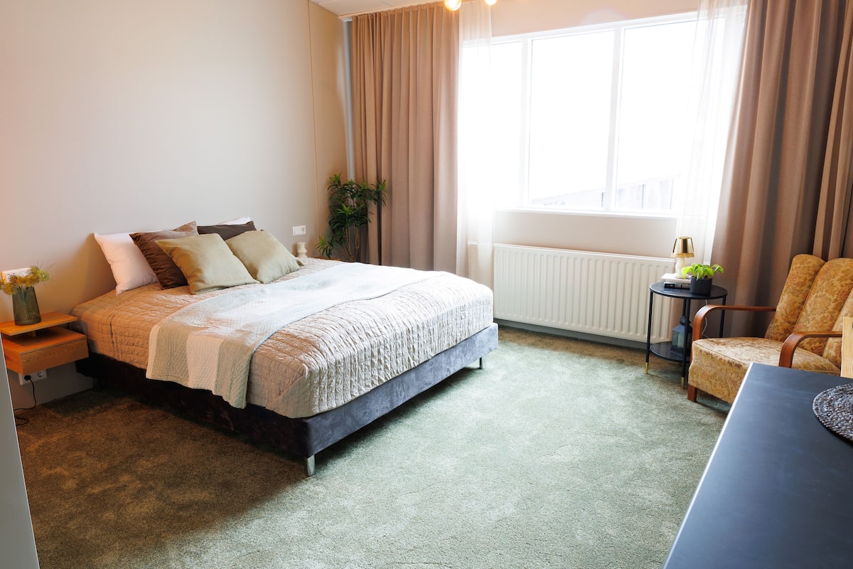 A bedroom with a bed, bedside table, chair, and a view of the outdoors through a window.