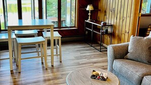 A cozy living room with a wooden floor, a round coffee table, a sofa, and a dining area with a table and chairs. - Lakefront year round home, gorgeous views, central location (Remsen)