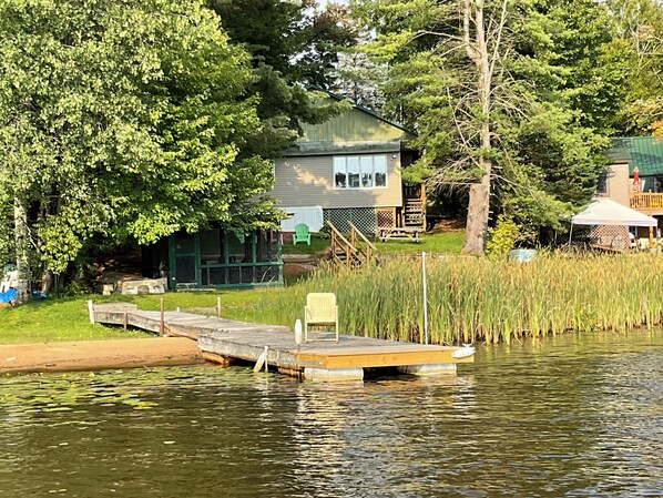 A dock extending into a calm body of water, surrounded by lush greenery and a house with a deck. - Lakefront year round home, gorgeous views, central location (Remsen)
