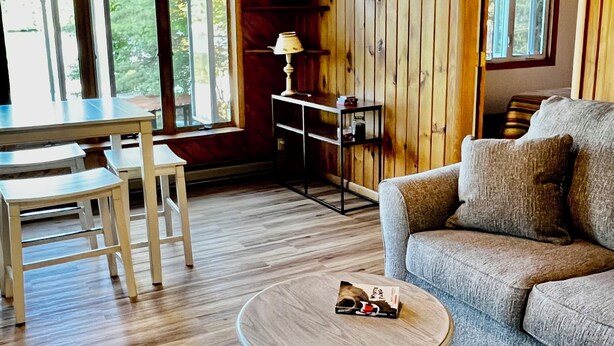 A cozy living room with a wooden floor, a round coffee table, a sofa, and a dining table with chairs. - Lakefront year round home, gorgeous views, central location (Remsen)