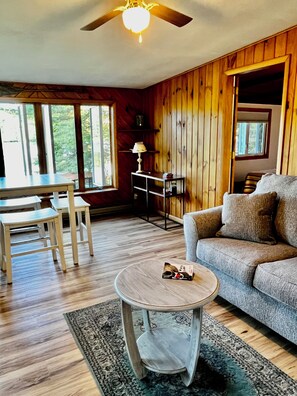 A cozy living room with a wooden floor, a round coffee table, a sofa, and a dining table with chairs. - Lakefront year round home, gorgeous views, central location (Remsen)
