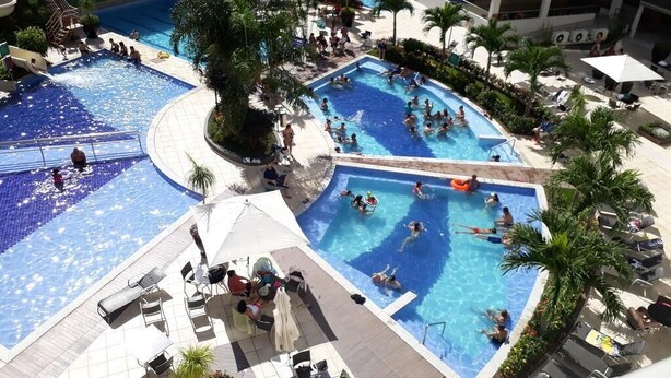 A rooftop pool area with multiple swimming pools, lounge chairs, and palm trees. - Rio Quente - Hotel Veredas do Rio Quente - Apartment for Rent (Rio Quente - Esplanada do Rio Quente)