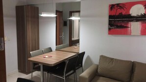 A modern living room with a wooden dining table, black chairs, a beige sofa, and a painting of a sunset over water. - Rio Quente - Hotel Veredas do Rio Quente - Apartment for Rent (Rio Quente - Esplanada do Rio Quente)