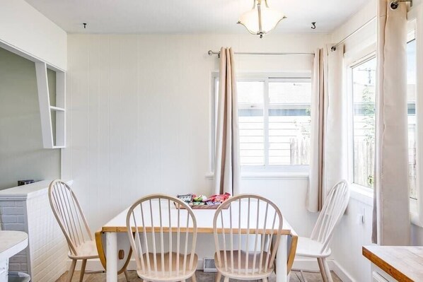 A dining area with a wooden table and four chairs, a window with curtains, and a white wall.