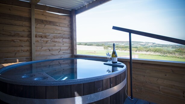 Eine Holzhütte mit Whirlpool, einer Weinflasche und einem Glas auf einem Tisch, mit Blick auf eine malerische Landschaft.