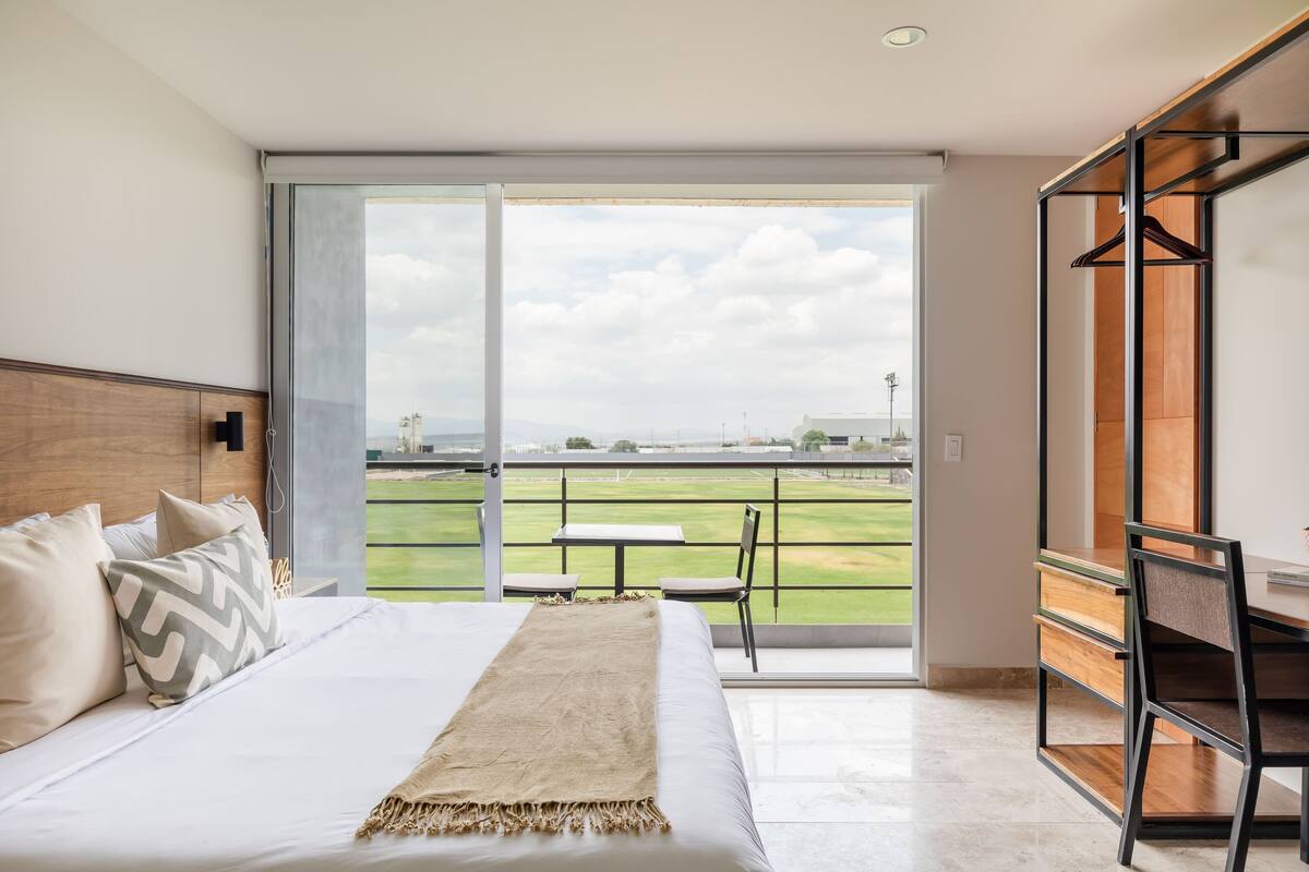 A hotel room with a bed, a desk, and a view of a grassy area through a large window.