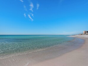 Een strand met helder turquoise water, een zandstrand en een paar gebouwen op de achtergrond.