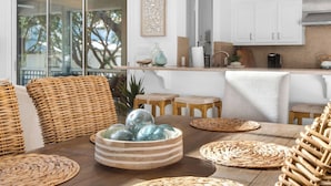 Una cocina moderna con armarios blancos, una mesa de comedor de madera, mobiliario de mimbre y un amplio ventanal con vistas a árboles.