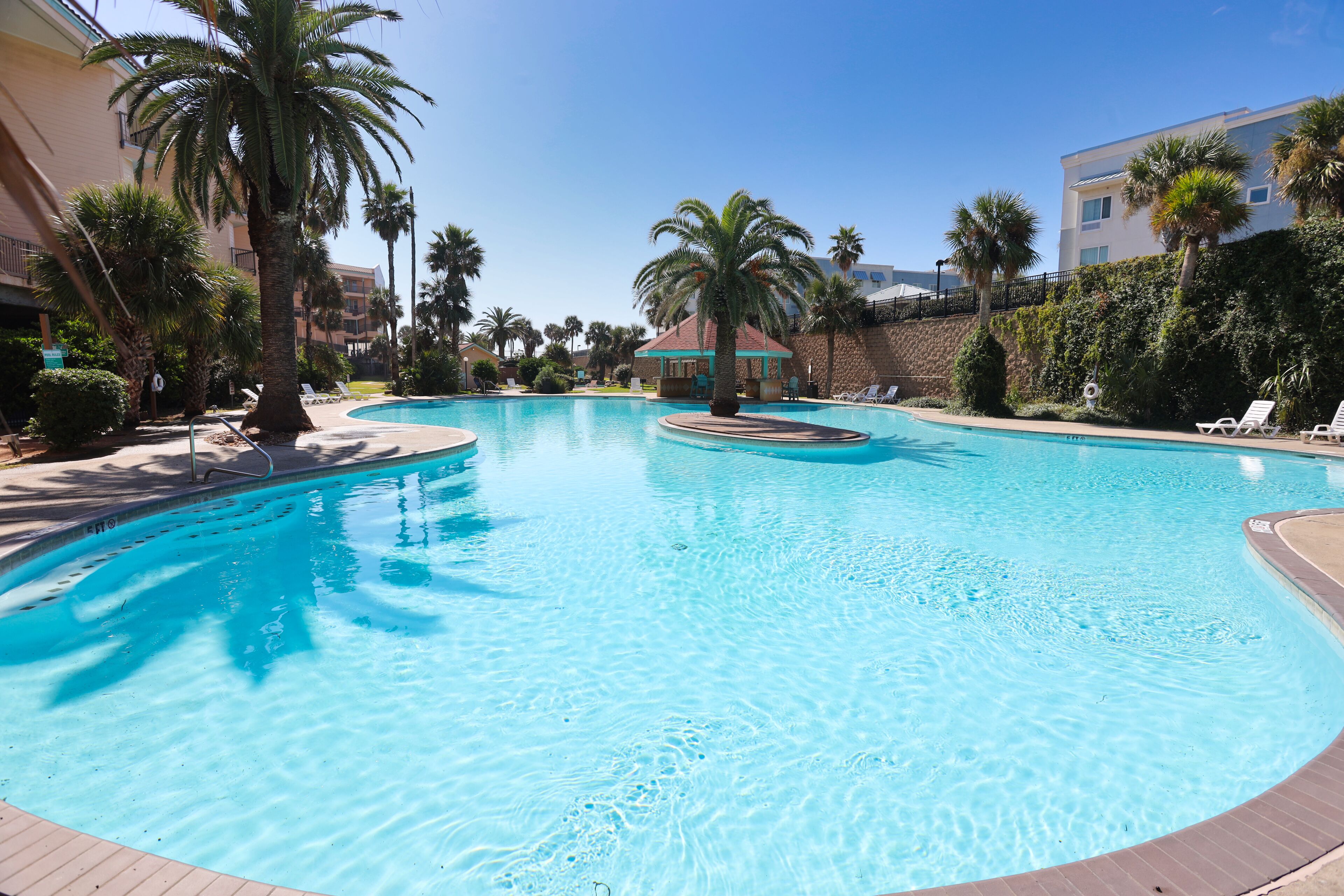 A swimming pool with palm trees, lounge chairs, and a building in the background.