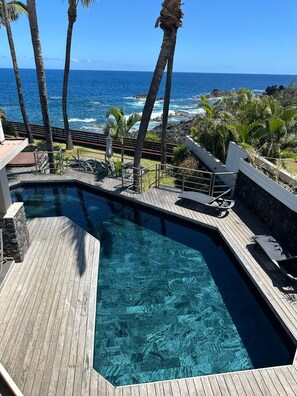 A clear blue pool with wooden deck, surrounded by palm trees and overlooking the ocean.