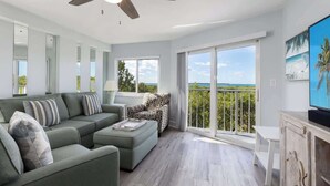 A modern living room with a sofa, ottoman, and patterned armchair. A flat-screen TV is on a wooden cabinet. Large windows offer a view of the ocean. - Spacious End Suite with Atlantic Views (Tavernier)