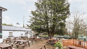 An outdoor seating area with wooden picnic tables and benches, surrounded by stone walls and a large tree. - Scotch Hall Cottage (Llangollen)