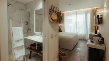 A modern hotel room with a shower, a sink, a bed, and a bedside table with a lamp.