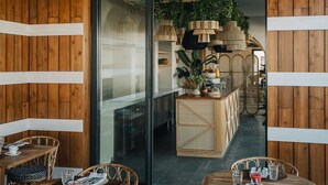 A wooden interior with tables and chairs, a glass door leading to a kitchen area, and hanging plants.