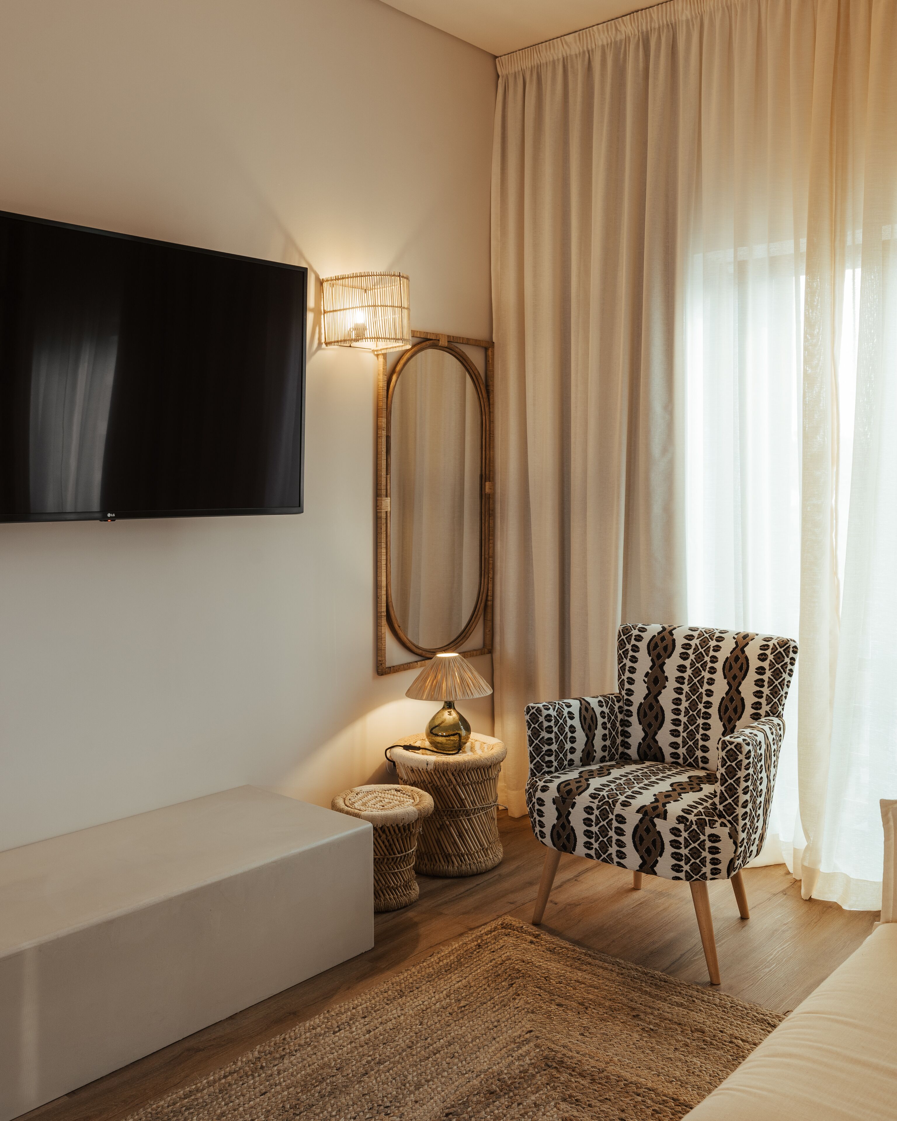 A modern living room with a flat-screen TV mounted on the wall, a patterned armchair, a round side table with a lamp, and a large mirror.