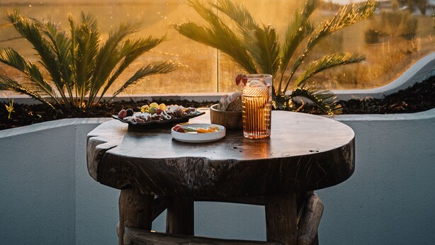 A table with a drink and snacks, overlooking a scenic landscape with trees and a sunset.