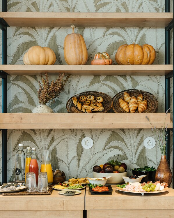 A buffet setup with various food items, including fruits, vegetables, and pastries, displayed on wooden shelves against a patterned wallpaper background.