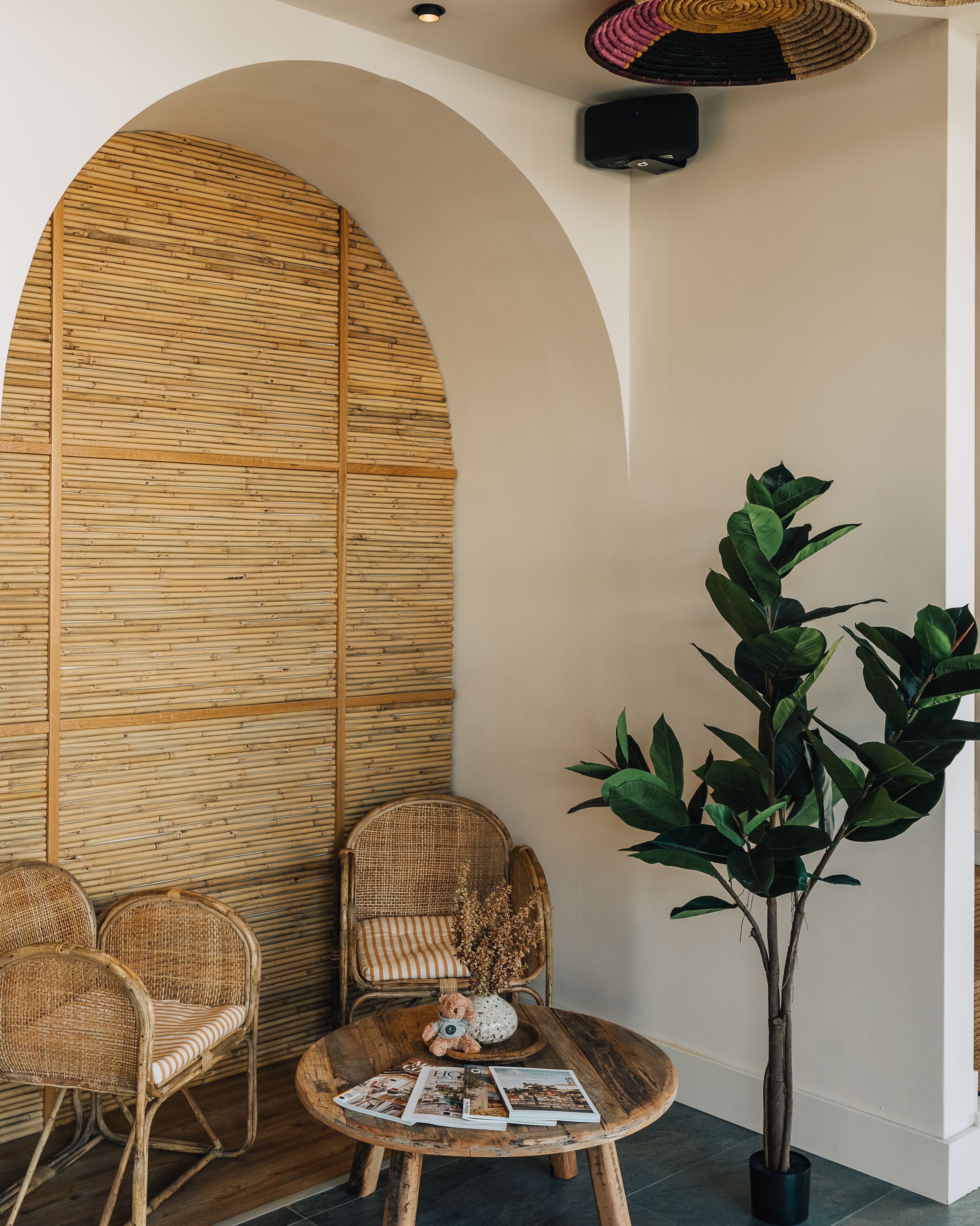 Ein Zimmer mit einer großen Holzwand, einem runden Holztisch mit Zeitschriften, zwei Korbstühlen, einer Zimmerpflanze und einer Deckenleuchte.
