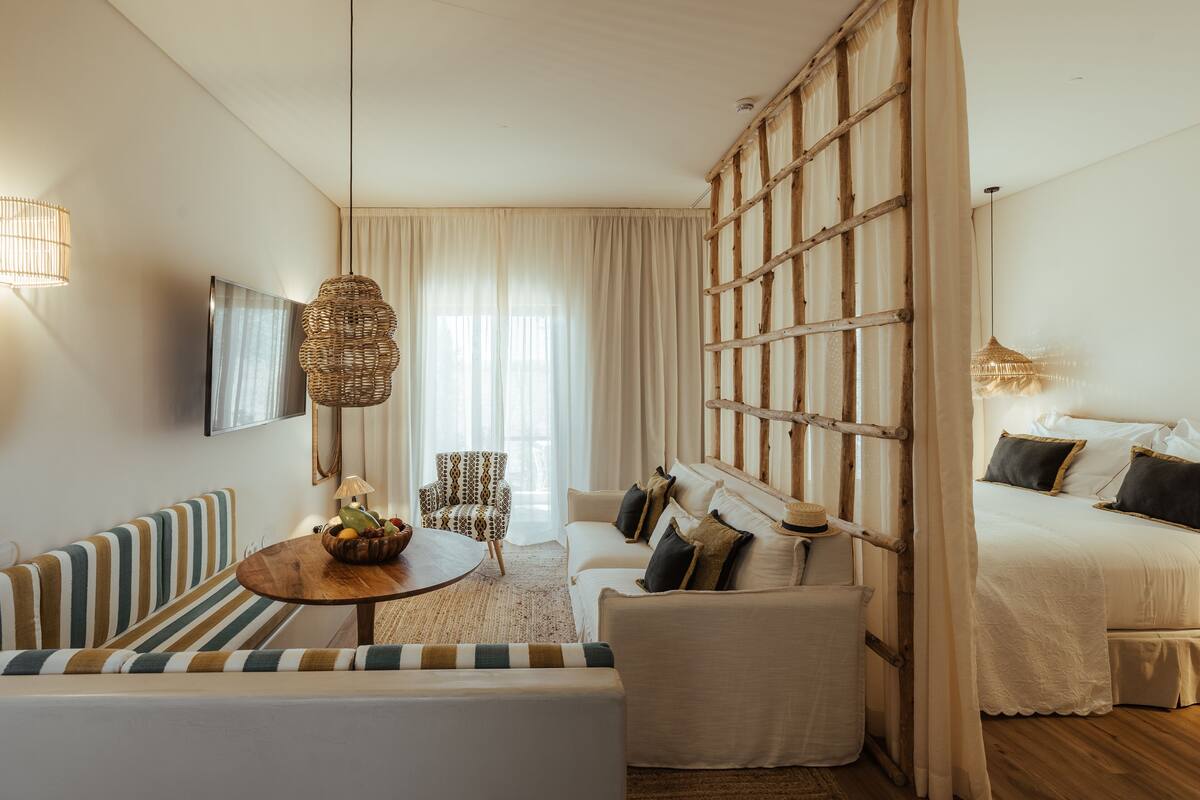 Ein modernes Hotelzimmer mit einer Couch, einem Esstisch, einem Bett und einem großen Fenster mit Vorhängen.