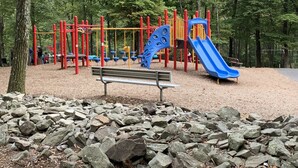 Ein Spielplatz mit einer blauen Rutsche und Klettergerüsten, umgeben von Bäumen und einem felsigen Vordergrund.