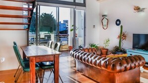 A modern living room with a wooden dining table, green chairs, a leather sofa, and a TV on a blue cabinet.