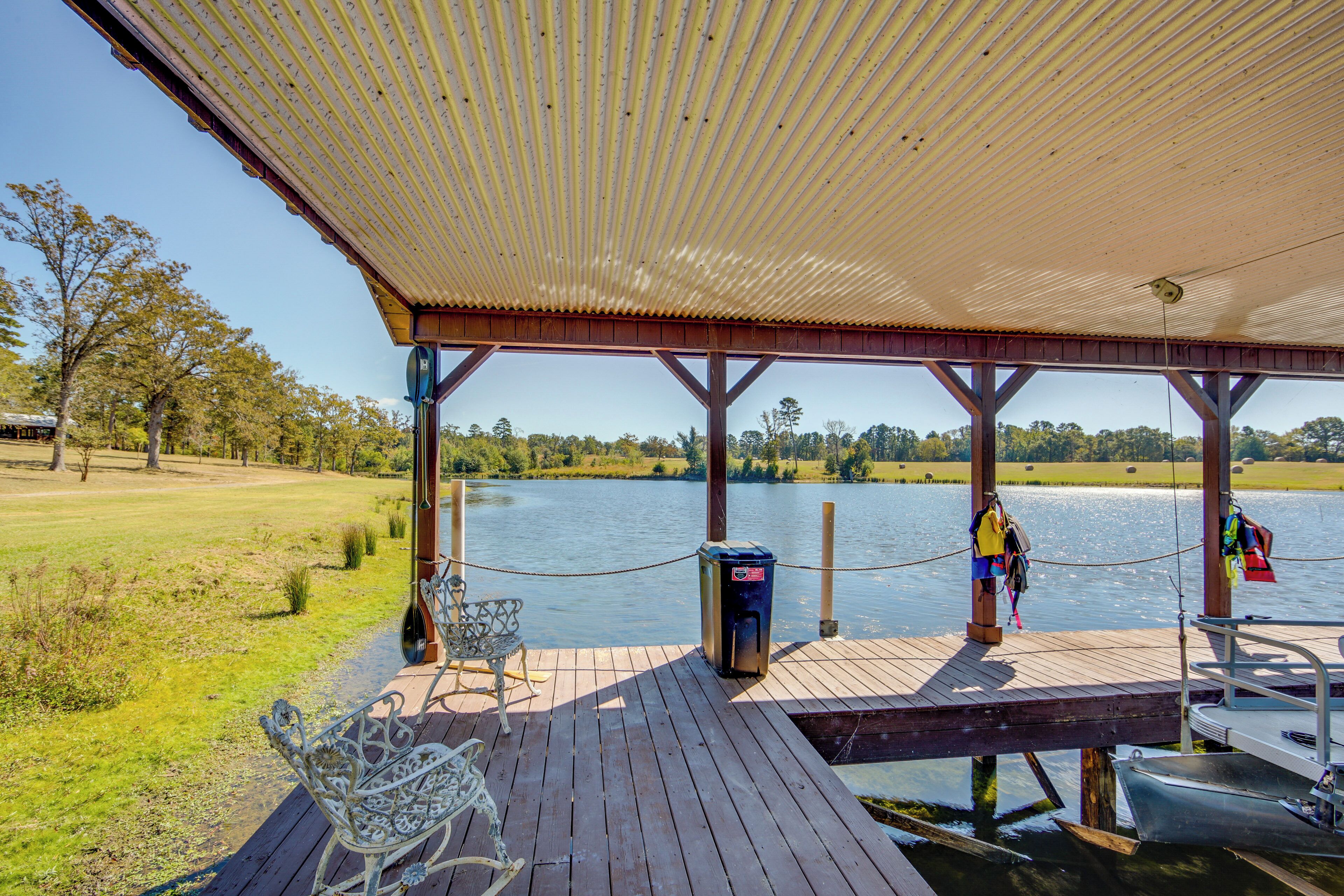 A wooden deck with a covered area overlooking a lake, a metal bench, and a small boat docked at the edge.