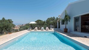A rectangular swimming pool with clear blue water, surrounded by a tiled deck. There are several lounge chairs and a white umbrella. A potted plant is visible on the right side. - Villa Helonaki (Corfu)