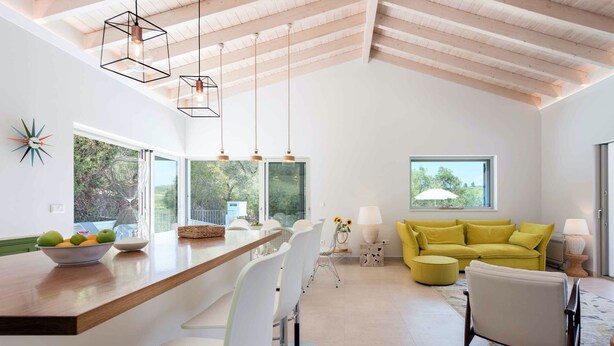 A modern kitchen with a central island, pendant lighting, and a view of the outdoors through large windows. - Villa Helonaki (Corfu)