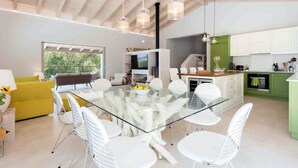 A modern kitchen with white cabinets, a dining area with a glass table and white chairs, and a large window with a view of greenery. - Villa Helonaki (Corfu)