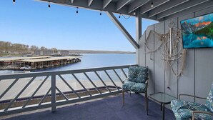 A balcony with a view of a dock and water, featuring two patterned chairs and a table, with a painting on the wall. - Turtle Haven - Relax & Enjoy the Incredible Views! (Osage Beach)