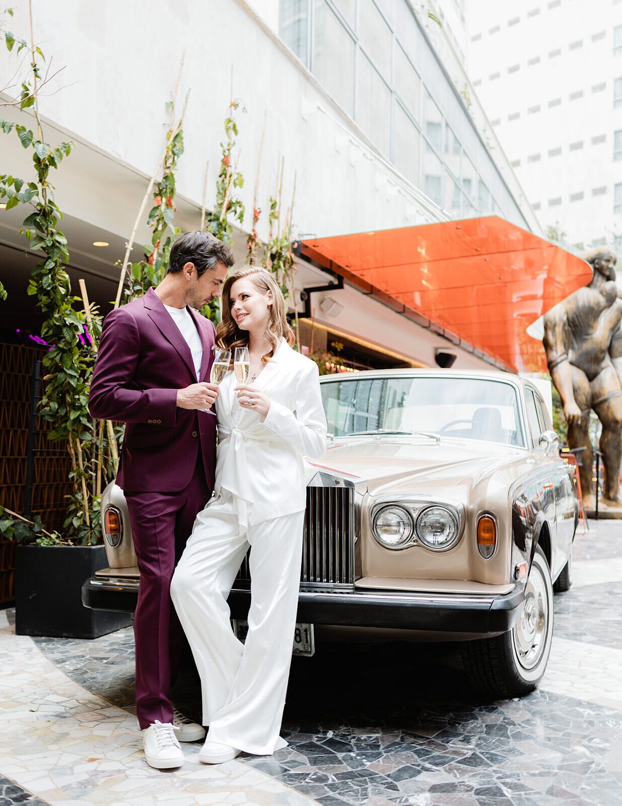 A couple standing next to a classic car with a modern building in the background.