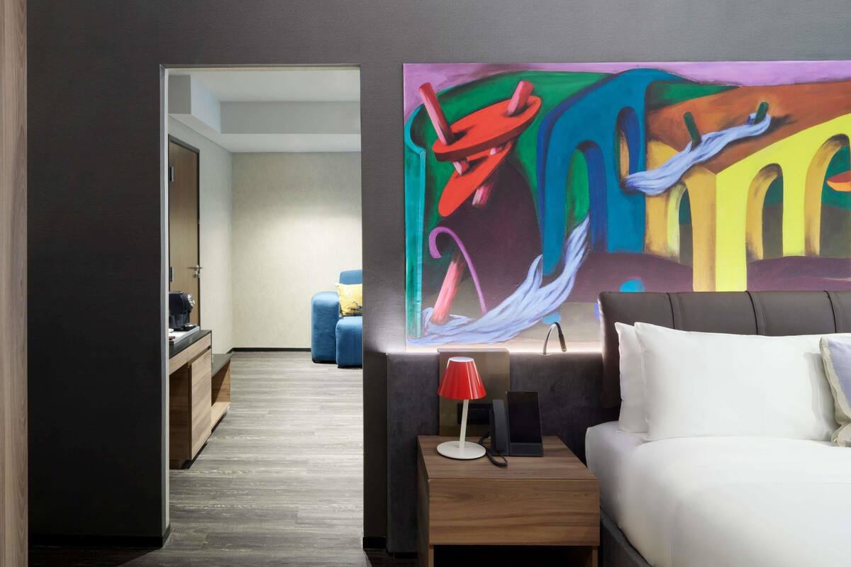 Een moderne hotelkamer met een groot abstract schilderij, een witte bank, een houten nachtkastje met een rode lamp en uitzicht op de hal.