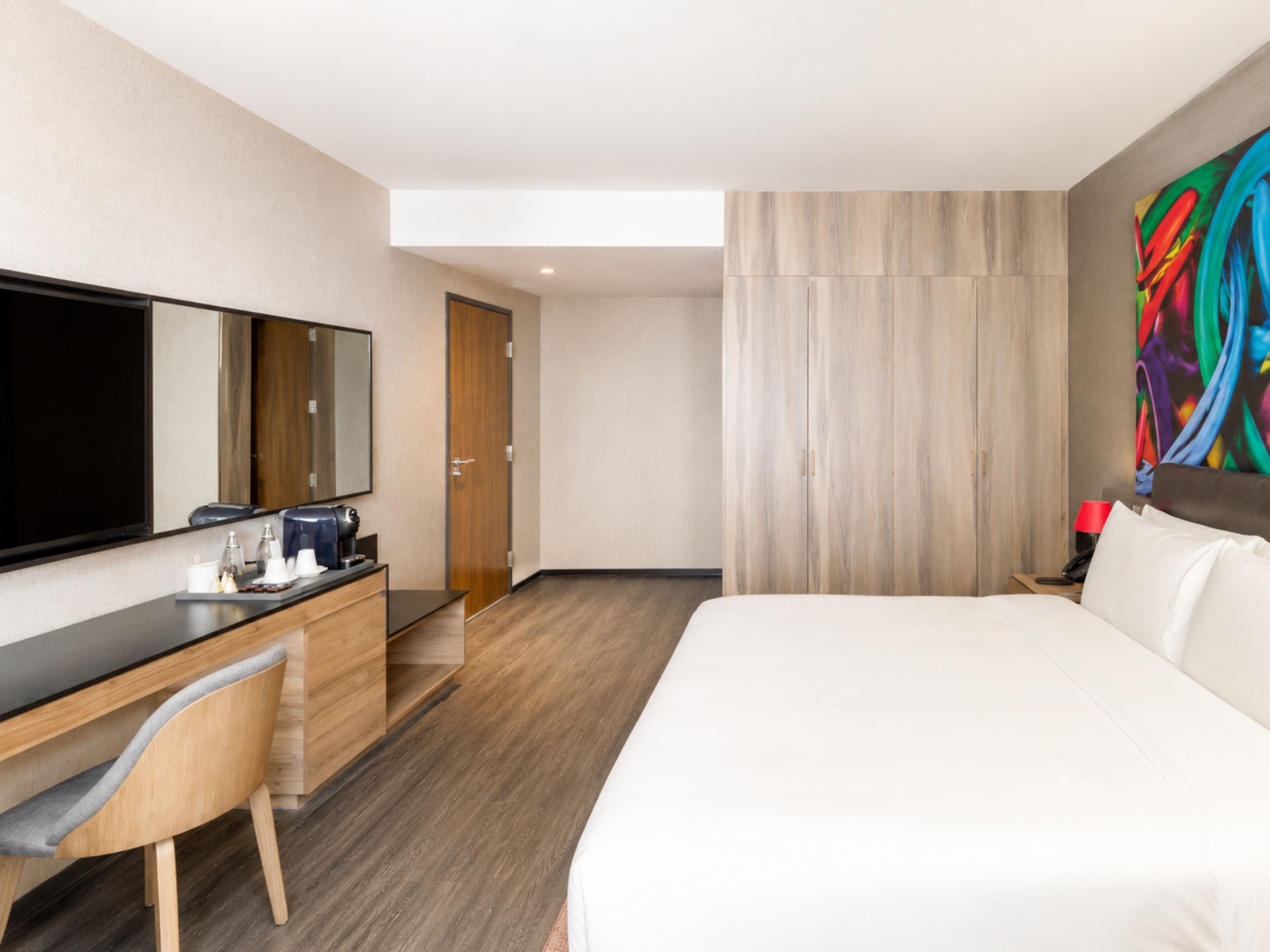 A hotel room with a bed, a desk with a chair, a television, a coffee machine, and a colorful abstract painting on the wall.