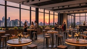 Un bar en la azotea con mesas y taburetes redondos, amplias ventanas con vista al horizonte de la ciudad y una barra con un bartender.