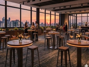 Un bar en la azotea con mesas y taburetes redondos, amplias ventanas con vista al horizonte de la ciudad y una barra con un bartender.