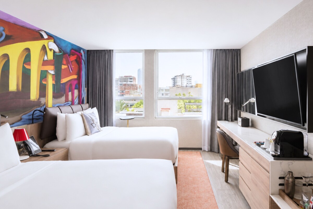 Habitación de hotel con dos camas, un escritorio, un televisor y un ventanal con vistas a la ciudad.
