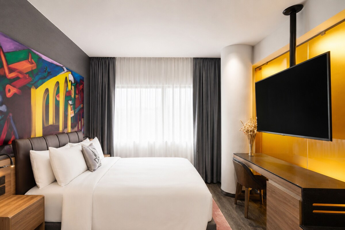 Una habitación de hotel moderna con una cama grande, un televisor de pantalla plana montado en la pared y un cuadro abstracto de colores en la pared.