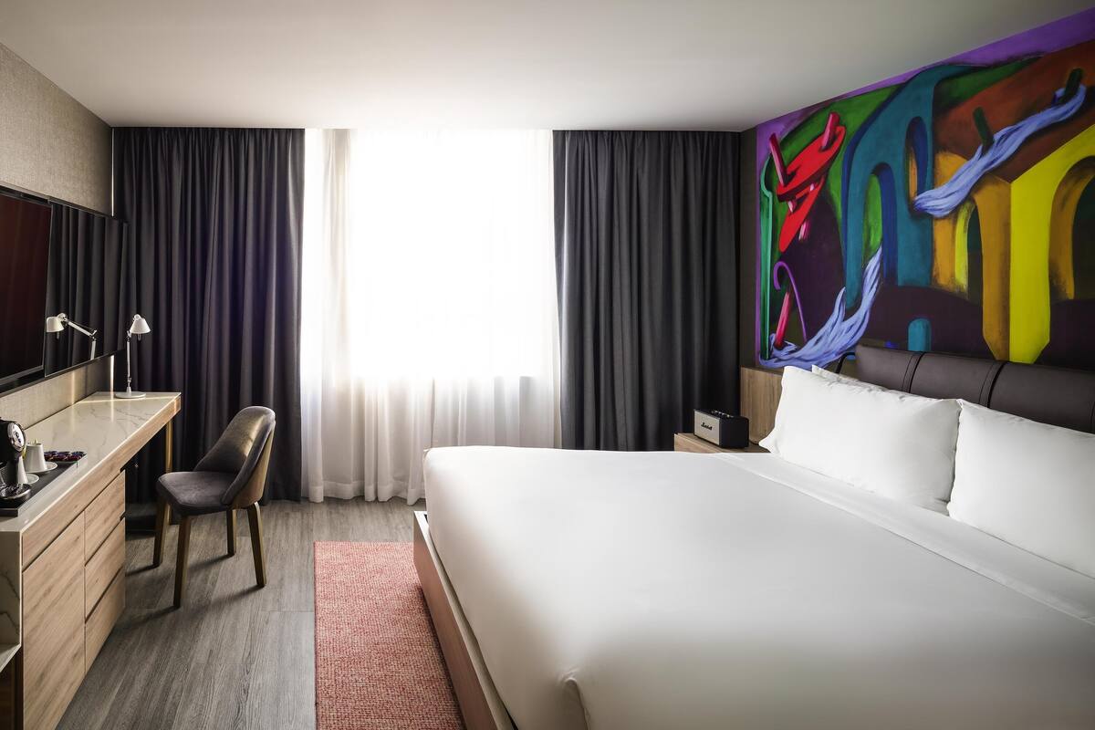 A hotel room with a bed, a desk, a chair, a TV, and a painting on the wall.